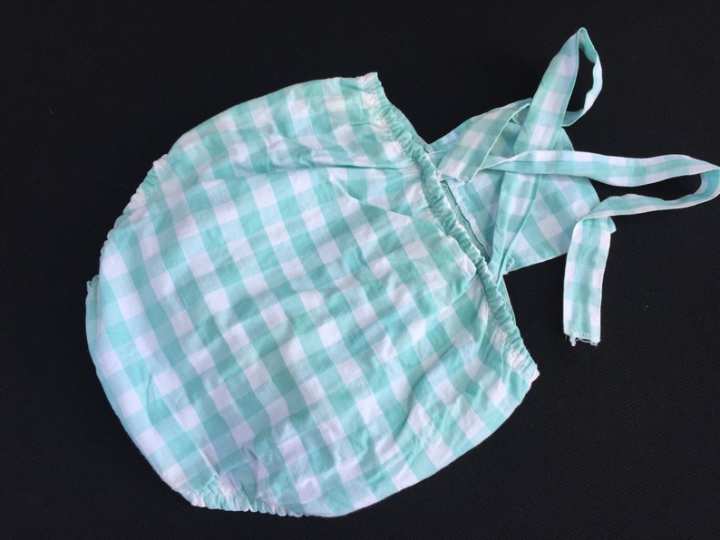 May include: A green and white gingham baby onesie with a matching strap. The onesie has a ruffled neckline and a gathered skirt.