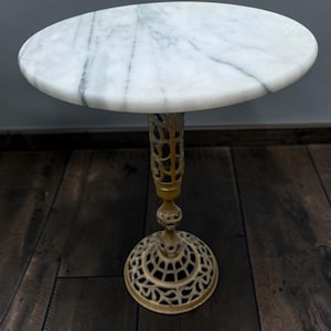 May include: A round side table featuring a white marble top with grey veining. The table is supported by a single, ornate, gold-toned metal pedestal base that features an intricate, openwork lattice design throughout the stem and the circular foot.