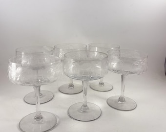 Könnte beinhalten: Ein Set aus sechs transparenten Champagner-Coupe-Gläsern. Jedes Glas hat eine runde Schale mit geätzten floralen Designs und einen schlanken Stiel. Die Gläser sind auf einer weißen Oberfläche angeordnet und zeigen ihr elegantes Design.