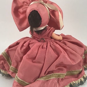 May include: A pink porcelain doll wearing a pink dress with a green trim and a large pink bonnet with a white ribbon.