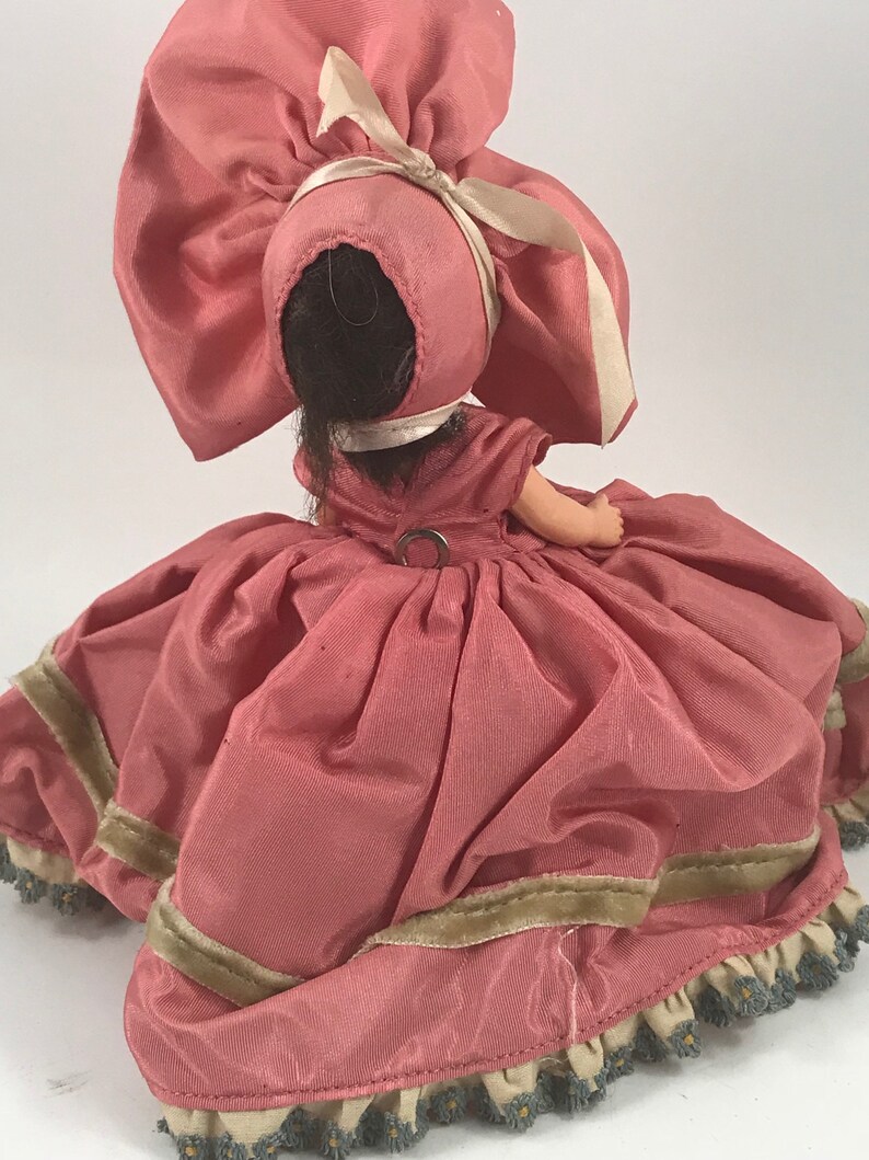 May include: A pink porcelain doll wearing a pink dress with a green trim and a large pink bonnet with a white ribbon.