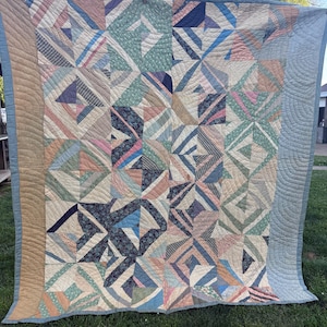 May include: A large, handmade quilt with a geometric pattern. The quilt features a patchwork design with various fabrics in shades of blue, green, pink, and beige. The border is a solid light brown, and the backing is a light blue.