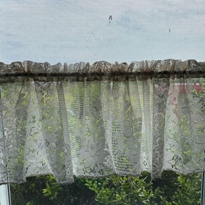 以下が含まれることがあります： 緑の葉が見える窓に、フリルのある白いレースのカーテンがかけられています。