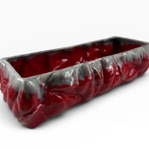 May include: A rectangular ceramic planter featuring a deep red glossy glaze with a textured, organic surface. The upper rim and edges are accented with a contrasting grey, drip-like glaze effect that highlights the uneven, sculpted exterior.