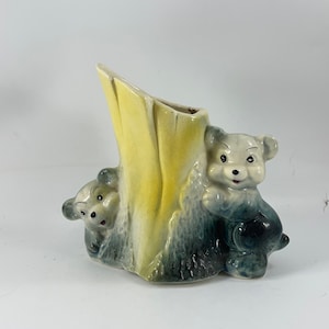 Puede incluir: Un macetero de cerámica con forma de tronco de árbol con dos osos asomando. El macetero está pintado en tonos amarillos, azules y grises.