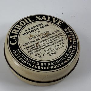 Könnte beinhalten: Eine weiße Dose mit schwarzer Beschriftung, die "CARBOIL SALVE" und "A SOOTHING ANTISEPTIC SALVE" für kleine Schnitte, Abschürfungen und Hautreizungen lautet. Die Dose ist mit "DISTRIBUTED BY NASHVILLE DRUG MFG. CO. 134 FOURTH AVENUE-NORTH-NASHVILLE, TENNESSEE" beschriftet.