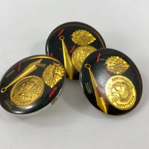 May include: Three gold and black round buttons with a design of a gold leaf, a gold spear, and a gold coin with a face on it. The buttons have a black background with red and gold accents.