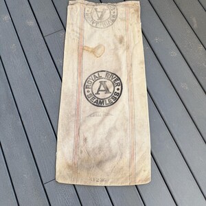 May include: A vintage, off-white canvas sack with red and blue stripes. The sack has a black circular stamp that reads "Royal River Seamless" with a large letter "A" in the center. The sack is labeled with the number "N1236".