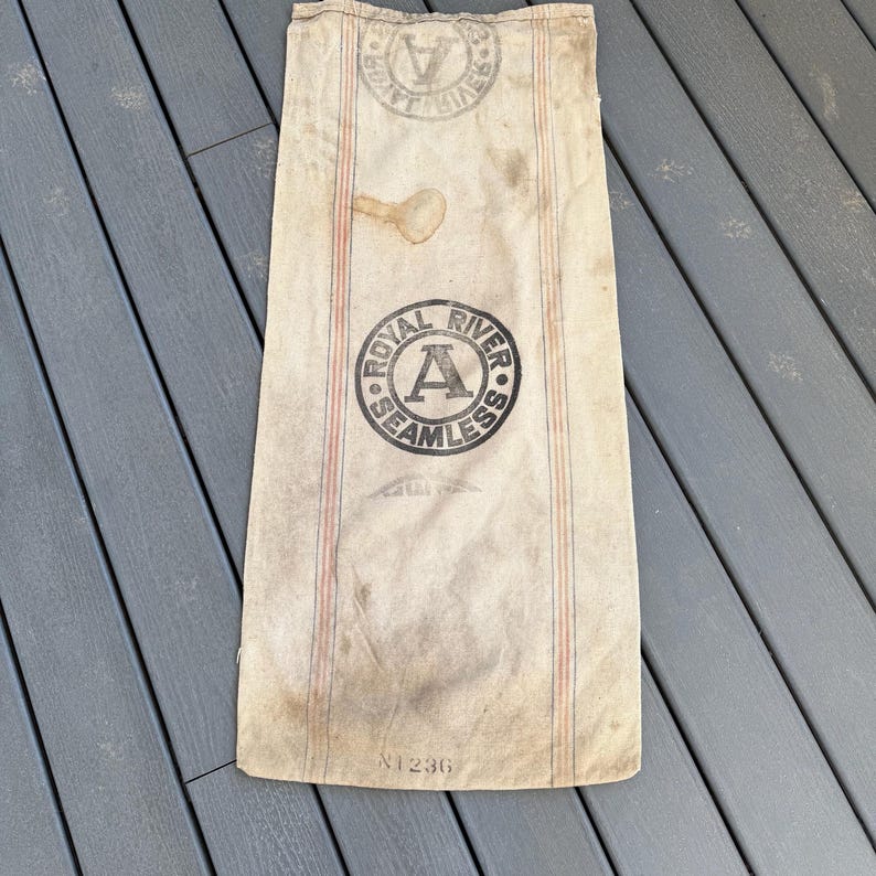 May include: A vintage, off-white canvas sack with red and blue stripes. The sack has a black circular stamp that reads "Royal River Seamless" with a large letter "A" in the center. The sack is labeled with the number "N1236".