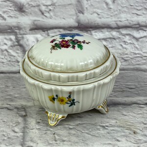 May include: A white porcelain trinket box with a lid, decorated with colorful floral designs and gold accents. The box has three gold-painted feet and a scalloped edge.