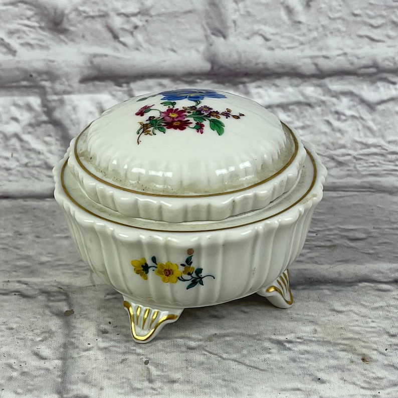 May include: A white porcelain trinket box with a lid, decorated with colorful floral designs and gold accents. The box has three gold-painted feet and a scalloped edge.