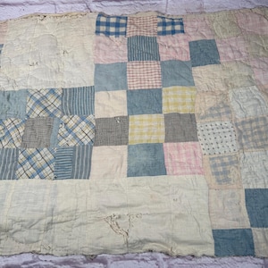 Puede incluir: Un edredón de retazos con una variedad de patrones y colores de tela. El edredón presenta cuadrados de telas azules, rosas, amarillas y blancas. Algunos cuadrados tienen patrones de cuadros o rayas. El edredón está sobre una superficie texturizada.