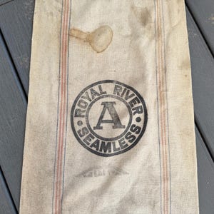 May include: A white canvas bag with red and blue stripes. The bag has a black circular stamp that says "Royal River Seamless" with a large letter "A" in the center.