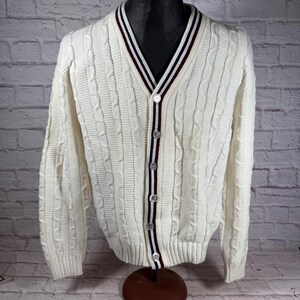 May include: A white cable knit cardigan with a v-neck and button closure. The cardigan has red, white, and blue stripes on the collar and down the front.