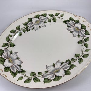 Puede incluir: Plato ovalado para servir con fondo blanco, con un diseño floral de flores blancas y grises con hojas verdes. El borde está adornado con dorado. Una pieza decorativa para servir comida.