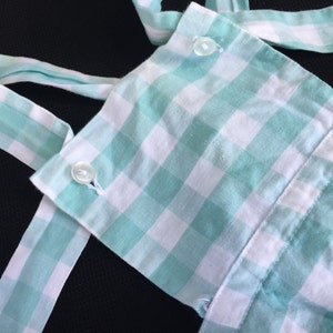 May include: A green and white gingham fabric with two white buttons. The fabric is folded and has a ruffled edge.