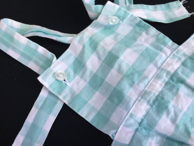 May include: A green and white gingham fabric with two white buttons. The fabric is folded and has a ruffled edge.