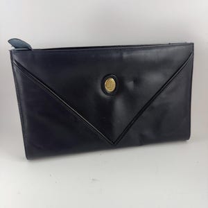 Peut inclure: Une pochette enveloppe en cuir noir avec un emblème circulaire doré. La pochette a une fermeture à rabat et une tirette en cuir. Le design présente un motif géométrique avec des lignes cousues.
