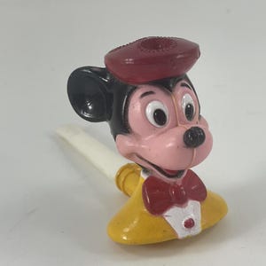 May include: A vintage Mickey Mouse figurine, featuring a red beret, black ears, and a pink face. The figure wears a yellow jacket with a red bow tie and a white collar. A white and gold-colored whistle extends from the figure's mouth.