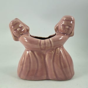 Puede incluir: Un jarrón de cerámica rosa vintage con dos figuras femeninas estilizadas abrazándose. Las cabezas de las figuras forman el borde del jarrón, con sus cuerpos fusionándose en una base común. El jarrón tiene un acabado brillante y un diseño clásico y elegante.