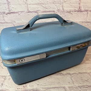 Puede incluir: Una maleta de viaje azul vintage con una superficie texturizada, detalles metálicos plateados y un asa azul a juego. La maleta tiene forma rectangular y un cierre de pestillo seguro. El asa mide unos 13 cm.