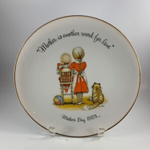 Könnte beinhalten: Ein weißer Keramikteller mit goldfarbenem Rand zeigt eine Vintage-Illustration von zwei Mädchen und einer Katze. Der Teller trägt den Text "Mother is another word for love" und "Mother's Day 1973..."