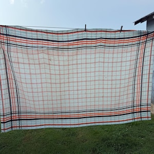 May include: A rectangular fabric cloth featuring a white base with a grid pattern of thin orange and black lines. The design includes wider horizontal and vertical bands of orange and black stripes along the edges, creating a classic plaid aesthetic.