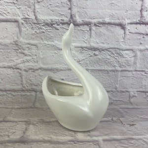 May include: A white ceramic swan planter with a curved neck and open basin. The swan is glossy and smooth, with a classic design. The planter is suitable for small plants or decorative items.