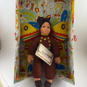 May include: A brown doll with a yellow butterfly costume and brown shoes. The doll is wearing a brown shirt with yellow polka dots and a brown hat. The doll is in a box with a colorful floral pattern.