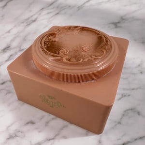 May include: A vintage peach-colored powder box with a decorative lid featuring floral and scrollwork designs. The box has a rectangular base and a round, ribbed lid. The text "Amy Lou" is visible on the front of the box.
