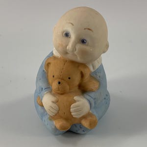 May include: A ceramic figurine of a baby in a blue outfit, holding a small brown teddy bear. The baby has a pale complexion, rosy cheeks, and blue eyes. The figurine is designed with a soft, rounded aesthetic.