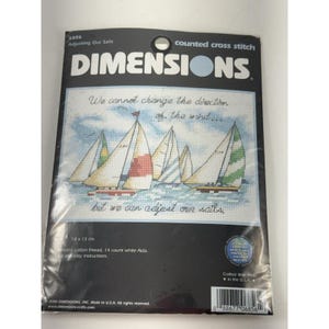 以下が含まれることがあります： Adjusting Our SailsというタイトルのDimensionsカウントクロスステッチキット。水上のヨットの航海シーンと、We cannot change the direction of the wind... but we can adjust our sailsというテキストが描かれています。パッケージのサイズは18 x 13 cmです。