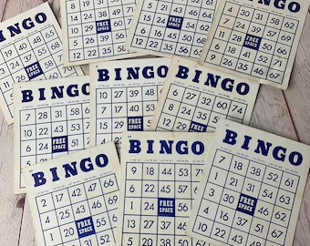 Può includere: Una collezione di nove carte da bingo vintage con testo blu e bianco su sfondo crema. Le carte sono disposte in modo casuale su una superficie di legno.