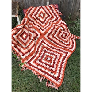 May include: A handmade crochet blanket featuring a repeating square pattern in shades of orange, red, cream, and white. The textile is constructed with a classic granny square design and is finished with a decorative fringe along the edges.