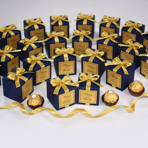 Può includere: Scatole regalo blu navy con fiocchi di raso dorati ed etichette personalizzate. Ogni scatola contiene un cioccolatino. Un nastro dorato è posto davanti alle scatole. Le scatole sono disposte su una superficie bianca.