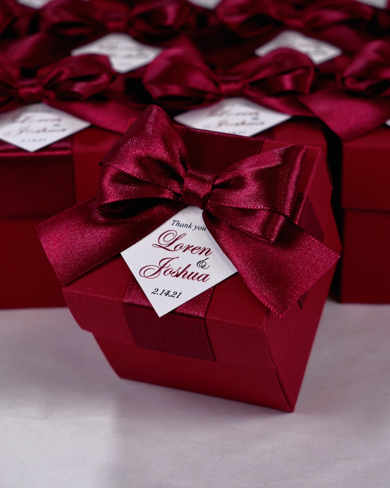 K&ouml;nnte beinhalten: Bordeauxfarbene Geschenkboxen mit einer Schleife aus Satinband und einem wei&szlig;en Etikett mit der Aufschrift "Danke Loren & Joshua 2.14.21".