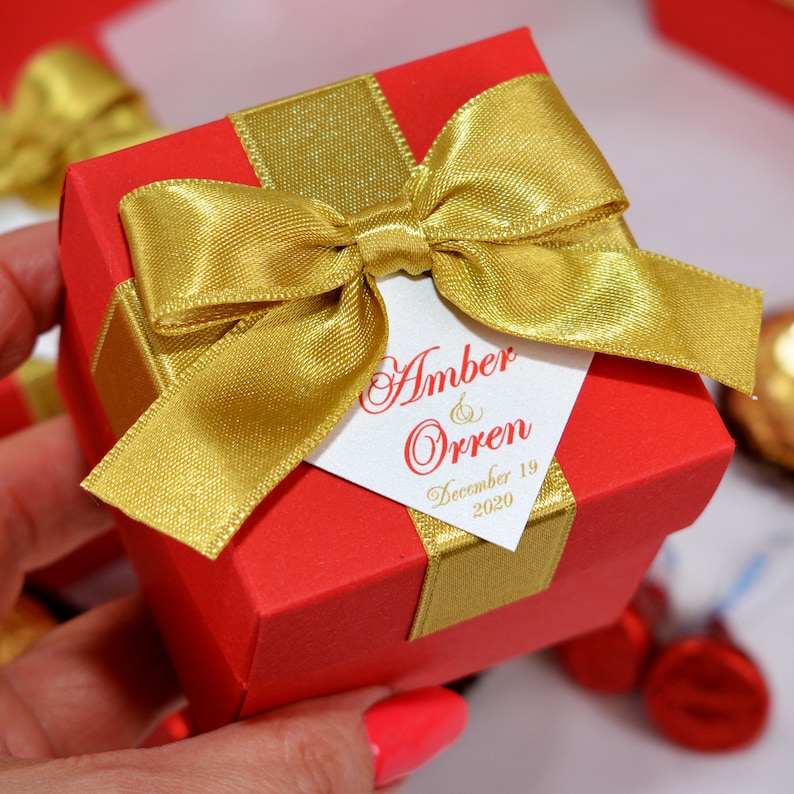 K&ouml;nnte beinhalten: Rote quadratische Geschenkbox mit goldener Schleife. Die Box hat ein wei&szlig;es Etikett mit dem Text "Amber & Orren December 19 2020".