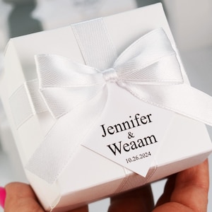 Pode incluir: Uma caixa de presente branca amarrada com uma fita e laço de cetim branco. Uma etiqueta branca com os nomes "Jennifer & Weaam" e a data "10.26.2024" está presa ao laço. A caixa é segurada por uma pessoa.