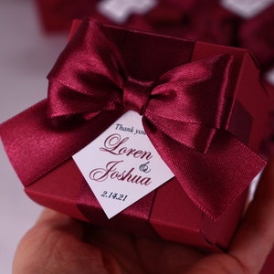 K&ouml;nnte beinhalten: Eine kleine, burgunderfarbene Geschenkbox mit einem Satinband, das zu einer Schleife gebunden ist. Die Box hat einen wei&szlig;en Anh&auml;nger mit dem Text "Thank you Loren & Joshua 2.14.21".