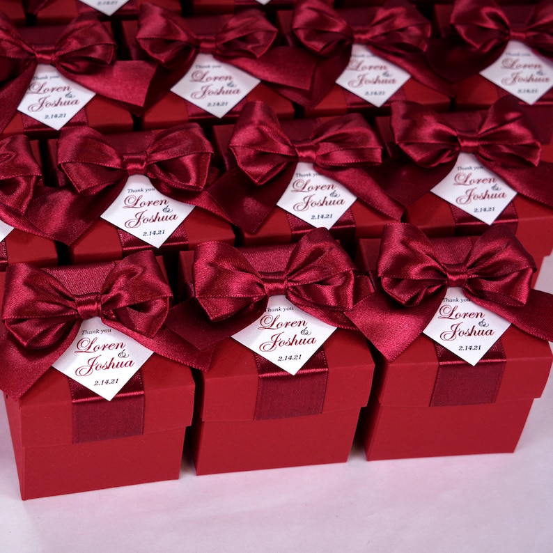 K&ouml;nnte beinhalten: Rote Geschenkboxen mit wei&szlig;en Satinb&auml;ndern und personalisierten Etiketten mit der Aufschrift "Danke Loren & Joshua 2.14.21".