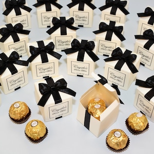 Può includere: Scatole regalo color avorio con fiocchi di raso neri, ciascuna con un'etichetta stampata. Sono visibili diversi cioccolatini dorati avvolti in carta stagnola. Le scatole sono disposte su una superficie chiara. Il testo sull'etichetta recita "Crystle's Sweet Memis 11.04.26".