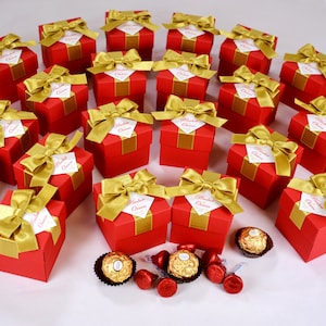 K&ouml;nnte beinhalten: Rote und goldene Geschenkboxen mit einer Schleife oben. Jede Box hat ein wei&szlig;es Etikett mit dem Text "Amber Orren" darauf gedruckt. Die Boxen sind in einem Kreis auf einer wei&szlig;en Oberfl&auml;che angeordnet. In der Mitte des Bildes befinden sich auch mehrere Schokoladenbonbons.