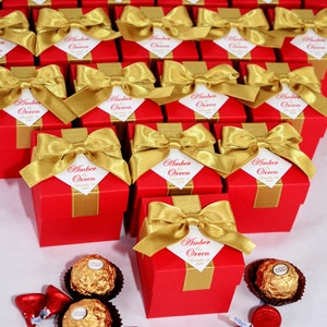 K&ouml;nnte beinhalten: Rote und goldene Geschenkboxen mit einem wei&szlig;en Etikett, auf dem "Amber & Orren" und das Datum "19. Dezember 2020" steht. Die Boxen sind mit einem goldenen Band und einer Schleife gebunden. Das Bild zeigt auch Schokoladenbonbons.