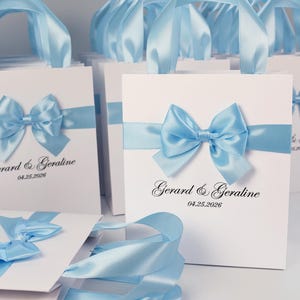 Könnte beinhalten: Weiße Geschenktüten mit hellblauen Satinbandgriffen und Schleifen. Die Taschen sind mit einem hellblauen Band und einer Schleife verziert und tragen die Namen "Gerard & Geraldine" und das Datum "04.25.2026".