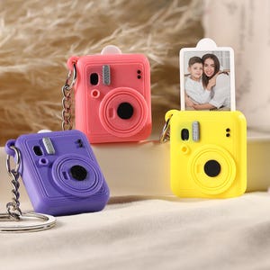 Peut inclure: Trois porte-clés miniatures en forme d'appareil photo, roses, violets et jaunes. L'appareil photo rose a une chaîne argentée et une petite photo. L'appareil photo violet a un porte-clés argenté. L'appareil photo jaune a une petite photo. Chaque appareil photo a un objectif circulaire.