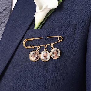 May include: A gold-toned safety pin brooch featuring three circular charms, each containing a small portrait photograph. The brooch is pinned to a navy blue suit jacket pocket, which also holds a single white calla lily flower and a grey necktie.