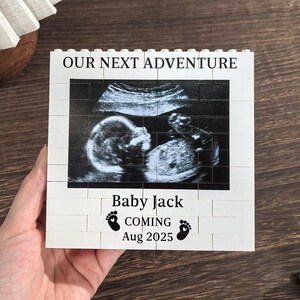 May include: A white brick-style block with the text "OUR NEXT ADVENTURE" at the top and a sonogram image in the center. Below the image, the text reads "Baby Jack COMING Aug 2025" with footprints. A pregnancy announcement.