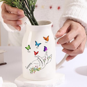 Könnte beinhalten: Eine weiße Keramikvase mit Henkel, verziert mit bunten Schmetterlingen und dem Wort "Granny" unter einer Handillustration. Die Vase enthält rosa Blumen und steht auf einem weißen Sockel.
