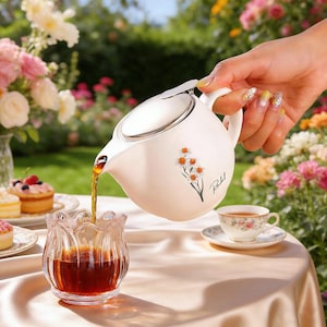 Könnte beinhalten: Eine cremefarbene Teekanne mit silbernem Deckel gießt Tee in eine klare Glas Tasse. Die Teekanne hat ein Blumendesign und die Initialen "RL" auf der Seite. Eine Tasse und Untertasse stehen auf dem Tisch, zusammen mit einem kleinen Kuchen. Der Hintergrund ist ein Blumengarten.