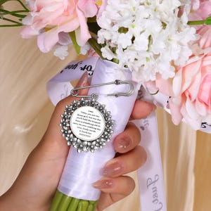 Puede incluir: Un broche floral plateado con un cierre de imperdible, sujeto a un ramo de flores rosas y blancas. El broche tiene un centro circular con texto grabado. El ramo está envuelto con una cinta blanca.